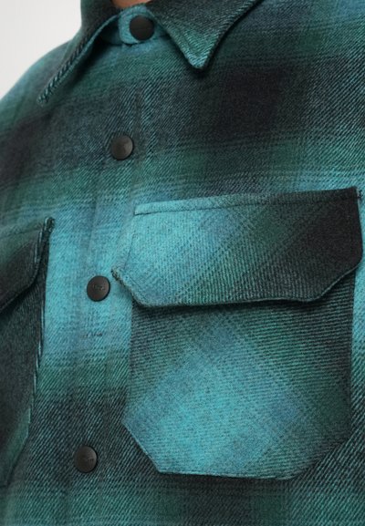 Camicia in flanella a quadri verdi e neri con due tasche sul petto e bottoni neri. Il materiale ha una finitura morbida e testurizzata.