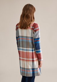 Vrouw met schouderlang bruin haar, gekleed in een lange geruite jas in rood, wit, blauw en beige, staand met haar rug naar de camera gekeerd tegen een effen achtergrond.