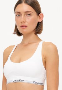 Bijela sportska grudnjak izrađen od glatkog materijala, s širokim trakama i logotip trakom s natpisom "Calvin Klein" u crnoj boji. Jednostavan, minimalistički dizajn.