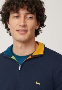 Maglione zip-up navy con accenti del colletto colorati (verde, arancione, giallo) e un piccolo logo di un cane giallo sul petto, tessuto a maglia strutturato.