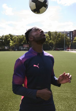 Man in marineblauwe en roze sportkleding die zich concentreert op een voetbal boven zijn hoofd op een zonnig buitenvoetbalveld.