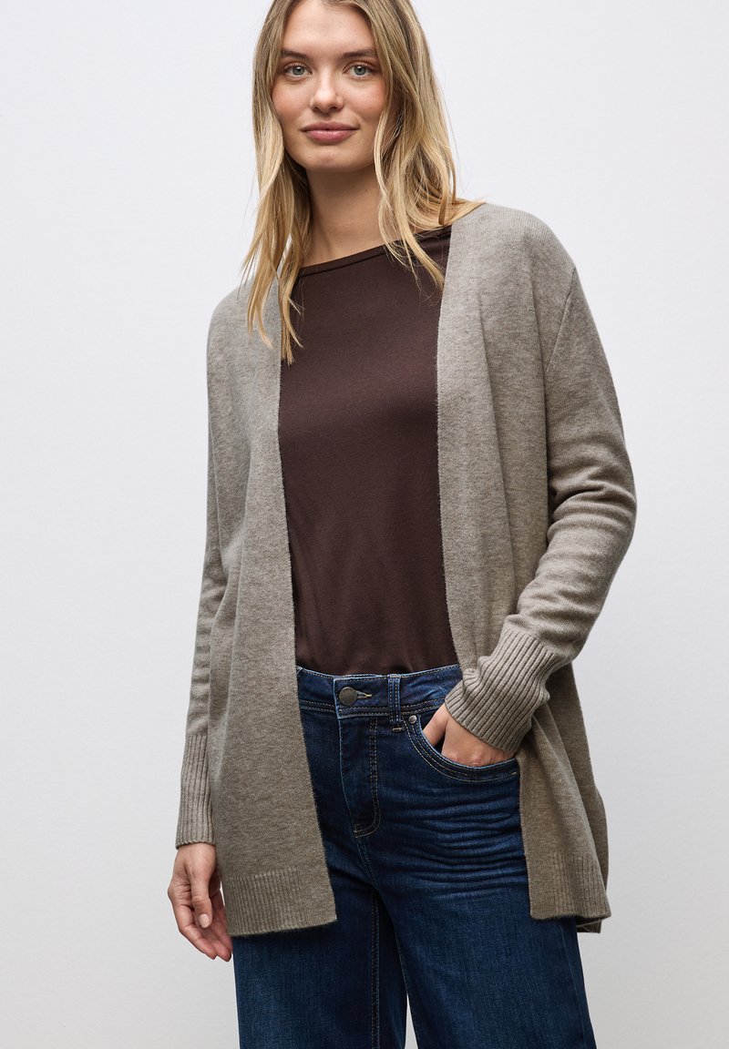 Jasnobrązowy kardigan z prążkowanymi mankietami i otwartym przodem, noszony na ciemnobrązowej bluzce, w połączeniu z niebieskimi dżinsami. Miękka tekstura, luzny krój.