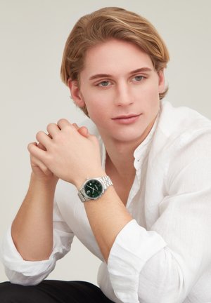Jeune homme en chemise blanche, reposant son menton sur ses mains jointes, portant une montre-bracelet argentée avec un cadran vert foncé, regardant légèrement sur le côté.