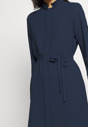 Marineblå kjole med høj krave og flæsekrave. Har lange ærmer og bindebælte i taljen for en figursyet silhuet. Blød tekstur.