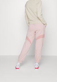 Osoba v světle růžových teplákách s tmavě růžovými akcenty a bílými teniskami Nike, stojící s rukama v kapsách, otočená zády.