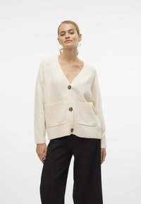 Cremefarvet strikket cardigan med V-udskæring, tre knapper og to forlommer. Har ribbede manchetter og kant for tekstur.