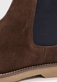 Braune Wildleder-Ankle-Boots mit einem navyblauen elastischen Einsatz, beiger Sohle und kontrastierenden Nähten entlang der Kante. Sanfte Textur und robuster Entwurf.