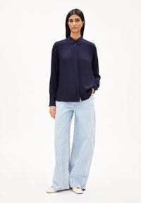 Navyblaue Satinbluse mit Kragen, langen Ärmeln und Knopfverschluss, kombiniert mit hellblauen weitem Jeans und cremfarbenen Schuhen.