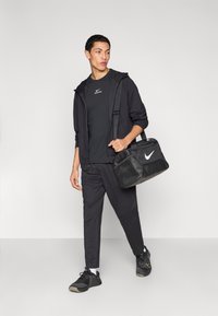 Μαύρο αθλητικό σύνολο που περιλαμβάνει φούτερ με φερμουάρ, μπλουζάκι και παντελόνι, σε συνδυασμό με μαύρη τσάντα γυμναστηρίου με λευκό λογότυπο και μαύρα αθλητικά παπούτσια.