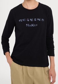 Persona che indossa una camicia nera a maniche lunghe con il testo in paillettes "WEEKEND Max Mara" abbinata a pantaloni beige, con una mano nella tasca.