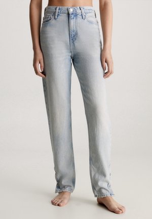 Personne portant un jean droit taille haute délavé clair avec des détails de boutons aux chevilles, debout pieds nus sur un fond uni.