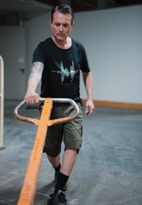 Camiseta negra con gráfico de onda sonora, pantalones cortos verdes oliva y zapatos negros, tirando de un carro de mano naranja en un entorno de almacén.