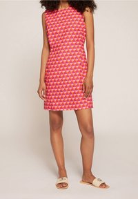 Rosa ärmelloses Kleid mit geometrischen Mustern in verschiedenen Schattierungen von Pink und Orange; kombiniert mit hellen, flachen Slipper-Sandalen.