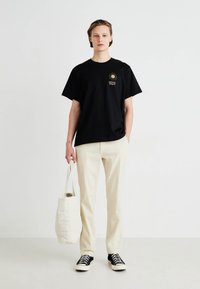 Tricou negru din bumbac cu un grafic aurit al soarelui și textul "Levi's", asortat cu pantaloni de culoare crem și adidași negri, ținând o geantă tote de culoare crem.