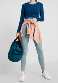 Langärmliges, marineblaues Oberteil mit blauem Kragen, hellrosa Jacke um die Taille gebunden, hellblaue Leggings, schwarze Sporttasche und weiße Turnschuhe.