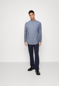 Camicia azzurra chiara con bottonatura frontale in tessuto lavorato, dotata di colletto classico e maniche lunghe, abbinata a pantaloni scuri e scarpe.