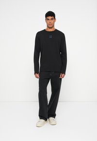 Vīrietis stāv pret baltu fonu, ģērbies melnā garā piedurknē kreklā, brīvās melnās bikses un baltos sporta apavus.