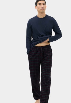 Mand iført navy langærmet skjorte og mørke ternede pyjamasbukser, løfter skjorten lidt for at vise maven, står barfodet på ensfarvet baggrund.