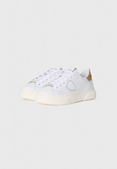 White sneakers with a textured upper, featuring a gray accent and a gold heel tab. Thick white outsole, traditional lace-up design.