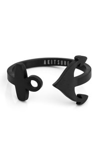 Anello in metallo nero con forme astratte, tra cui un segno più e una freccia stilizzata. Superficie liscia e design minimalista.
