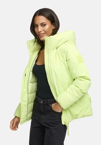 Vrouw in een lichtgroene pufferjack met capuchon, zwarte top en donkere jeans, lachend met één hand in de zak tegen een effen achtergrond.