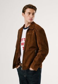 Giacca in suede marrone con colletto, chiusura con zip e tasche laterali. Indossata sopra una maglietta bianca con scritte rosse. I jeans di denim completano l'outfit.