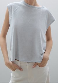 Valkoinen harmaa hihaton paita, jossa on pyöreä kaula-aukko, pehmeä rakenne, rento istuvuus ja pudotetut olkaimet. Yhdistetty vaaleanvärisiin housuihin.