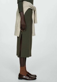 Robe mi-longue olive avec fente latérale, pull beige noué à la taille, chaussettes couleur rouille et mocassins en cuir marron foncé sur une personne debout.