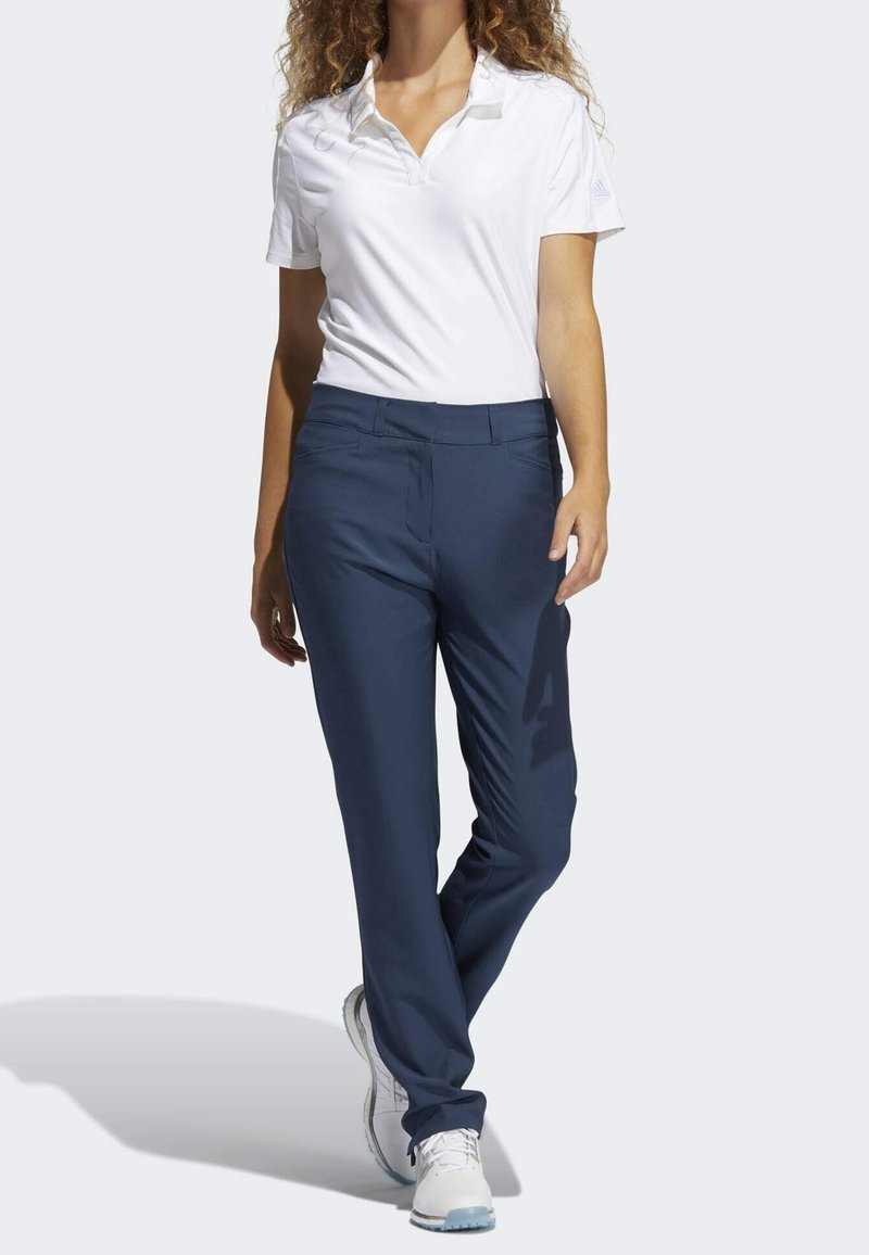Femme marchant portant un polo blanc à manches courtes, pantalon bleu marine et baskets blanches sur un fond uni.