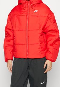 Röd puffjacka med huva, med en naturlig lyster, vadderad textur och en vit Nike-logotyp på vänster bröst. Dragkedjeslclosure framtill.