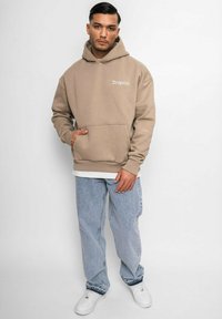 Brauner Kapuzenpullover mit Vorderntasche, lockere Passform. Kombiniert mit hellblauen Jeans und weißen Sneakers. Besitzt gestickten Text auf der Brust.
