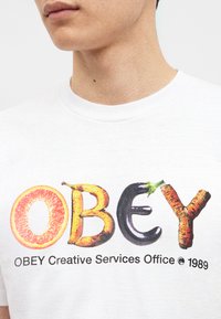 Κοντινό σε ένα λευκό μπλουζάκι με τη λέξη OBEY γραμμένη με εικόνες φρούτων και λαχανικών πάνω από το κείμενο "OBEY Creative Services Office 1989".