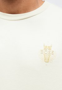 Ανοιχτό μπεζ κοντομάνικη μπλούζα, με ανάγλυφο χρυσό λογότυπο "BOSS" διακοσμημένο με στοιχεία, στρογγυλή λαιμόκοψη και απαλό ύφασμα.