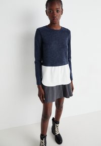 Pull en tricot marine à manches longues sur une chemise blanche à boutons et une jupe grise évasée ; associé à des bottines à lacets noires.