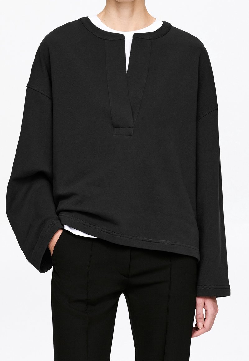 Personne portant un haut noir ample à manches longues avec une encolure en V profonde par-dessus une chemise blanche, associé à un pantalon noir ajusté, une main dans la poche.