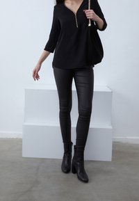 Donna che indossa un top nero con zip, pantaloni in pelle neri, stivaletti neri con tacco a caviglia e porta una borsa nera con tracolla in oro.