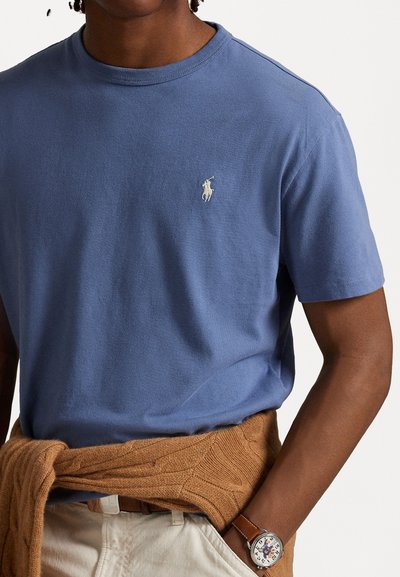 Blå bomulls t-shirt med rund hals och korta ärmar, med en liten vit logotyp på bröstet. Brun stickad tröja knuten runt midjan.