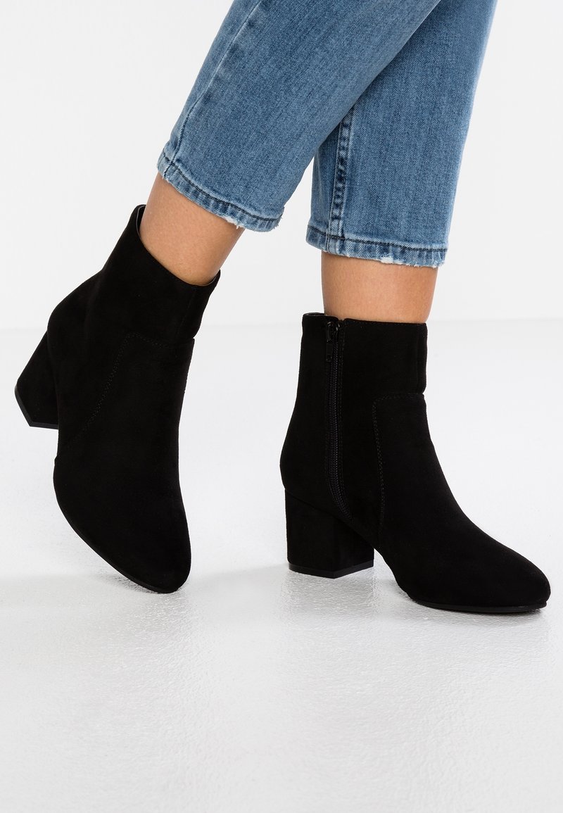 Personne portant des bottines en daim noir avec des talons blocs et des fermetures éclair latérales, associées à un jean denim bleu court sur fond blanc.