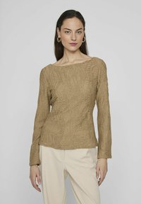 Top lungo a maniche lunghe di colore marrone con una scollatura ampia, caratterizzato da un motivo ondulato. Abbinato a pantaloni beige chiaro, il modello si trova davanti a uno sfondo neutro.