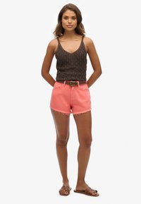 Top de punto marrón sin mangas y shorts de mezclilla rosa con dobladillo deshilachado, complementados con un cinturón marrón. Sandalias de tiras anchas completan el look.