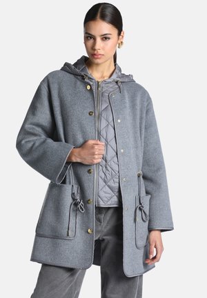 Femme portant une tenue grise en couches, comprenant une veste matelassée zippée avec capuche et un manteau en laine avec de grandes poches et des boutons dorés.