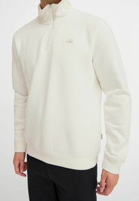 Sweatshirt crème clair en pull à col montant et zip mi-long ; il présente des poignets et un ourlet élastiques, avec un petit logo brodé sur la poitrine.