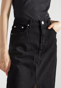 Gonna di jeans nera con spacco frontale, design a cinque tasche e dettagli in metallo argentato. La texture appare leggermente sbiadita.