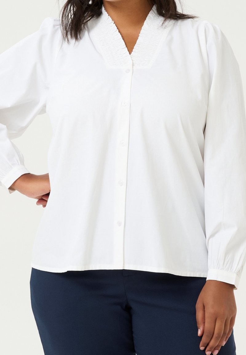 Femme portant un chemisier blanc à manches longues avec un col froncé et un pantalon bleu marine, debout avec une main sur la hanche.