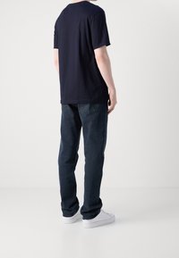 Camiseta azul marino de manga corta con una costura trasera sutil, combinada con jeans de mezclilla oscuros y zapatillas blancas, vista desde la parte trasera.