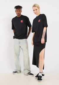 To modeller står side om side. Den ene bærer en sort oversized t-shirt, lyseblå jeans og sorte sneakers. Den anden bærer en sort spidsskjørt og t-shirt.