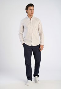 Va shirtipuseri pitkähihaisen nappipaidan, jossa on beige ja valkoinen pystysuora raita, yhdistettynä tummansinisiin housuihin ja valkoisiin lenkkareihin. Sileä kangas.