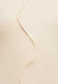 Μπεζ ριμπ κορυφή με κυματιστό σχέδιο, με μικρά στρογγυλά κουμπιά στο μπροστινό μέρος. Μαλακή υφή με εφαρμοστή σιλουέτα.