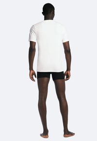 Hvid t-shirt med korte ærmer, tætsiddende design. Modellen bærer sorte shorts, står barfodet og viser det glatte stof og den enkle halsudskæring. Bagfra.