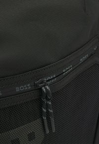 Czarny plecak z teksturowanej tkaniny, z siateczkową kieszenią, posiadający wyraźny suwak z logo w powtarzającym się wzorze.